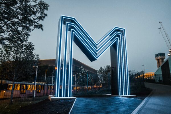 A large-scale steel structure of the letter M, illuminated at dusk